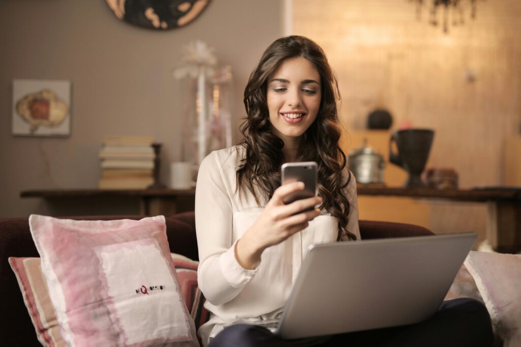 A woman using her phone and laptop while sitting on a couch