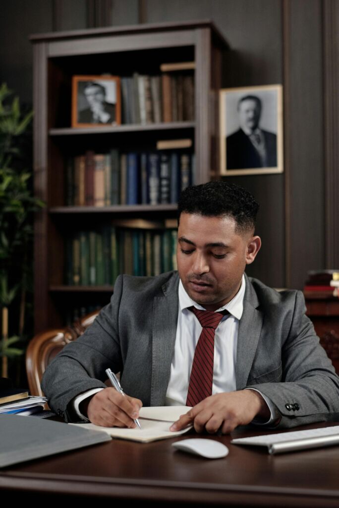 A man in a suit writing at a desk in a wood-paneled office