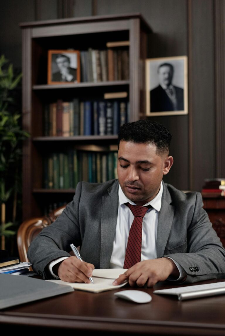 A man in a suit writing at a desk in a wood-paneled office